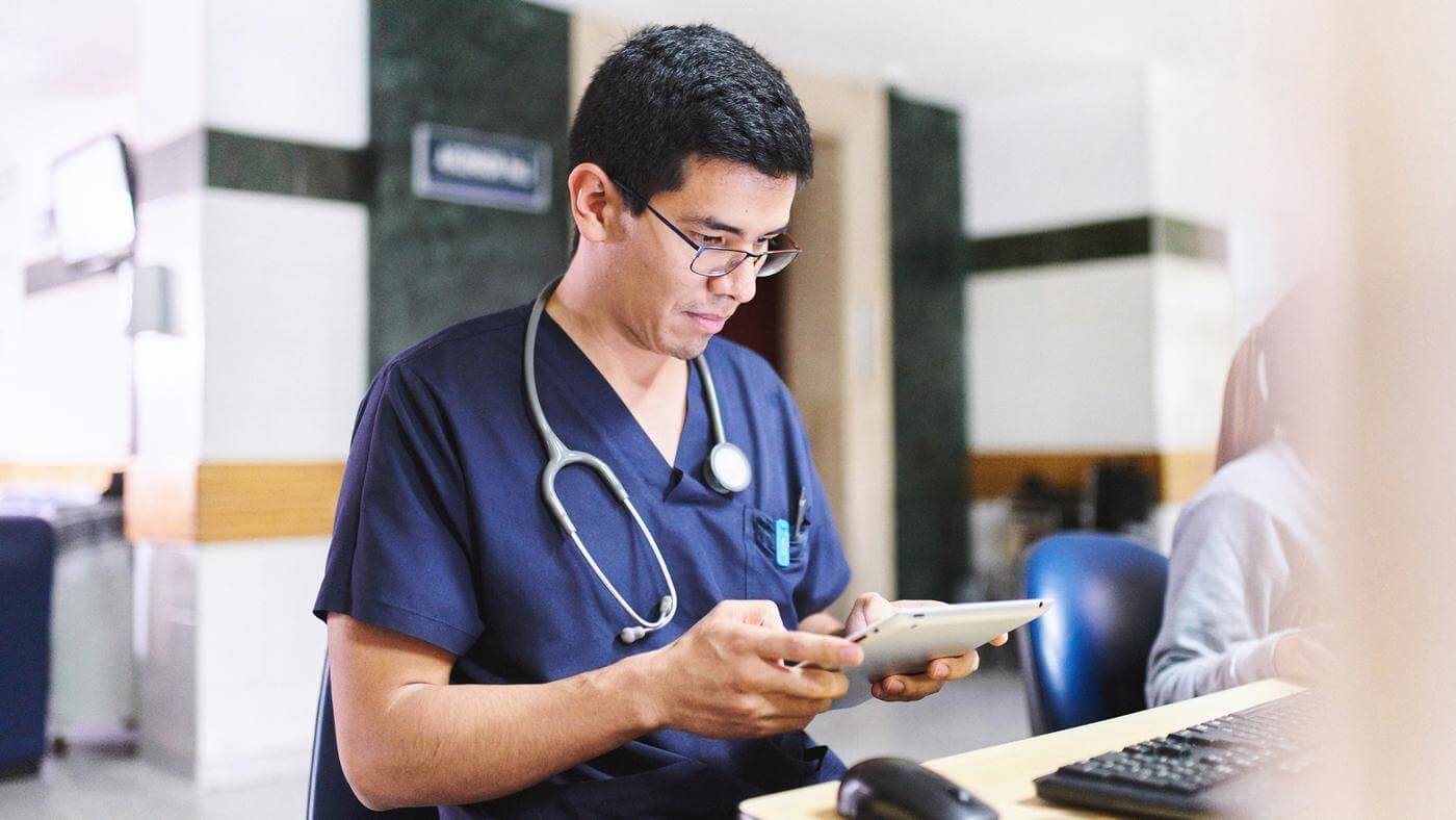 A male physician in scrubs is interacting with a tablet, likely reviewing patient information.