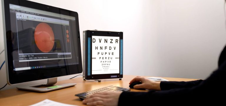 Person uses a computer during an eye examination, with a retinal image displayed on screen beside an illuminated vision chart.