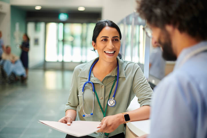 Healthcare professional with a stethoscope hands over paperwork at a reception desk inside a healthcare facility.